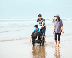 family pushes wheelchair on sandy beach as the teen in the wheelchair reaches for the wet sand.