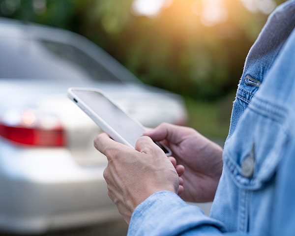 Person holding a smartphone near a parked car, wearing a denim jacket.