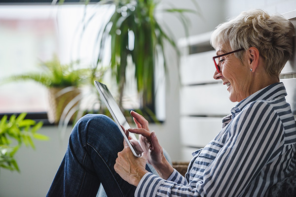 Older woman wearing glasses and a striped shirt, sitting by a window, smiling while using a tablet.