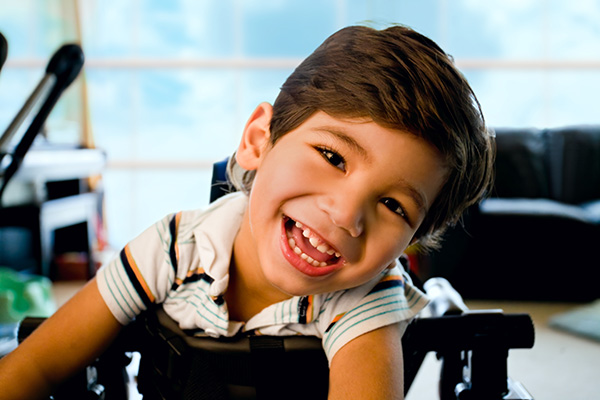 A young child with dark hair smiles while leaning forward in a wheelchair. The room has a bright, open background.