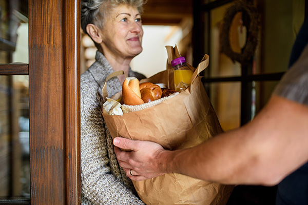 A person hands a paper bag filled with groceries, including bread and juice, to an older woman standing in a doorway.