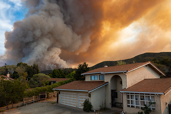 Smoke billows over hills behind a house under a cloudy sky, indicating a wildfire in the distance.