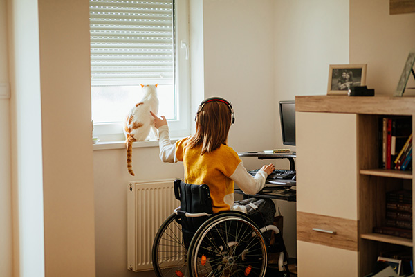 A person in a wheelchair wearing headphones sits at a desk with a computer, gesturing toward a cat on a windowsill.