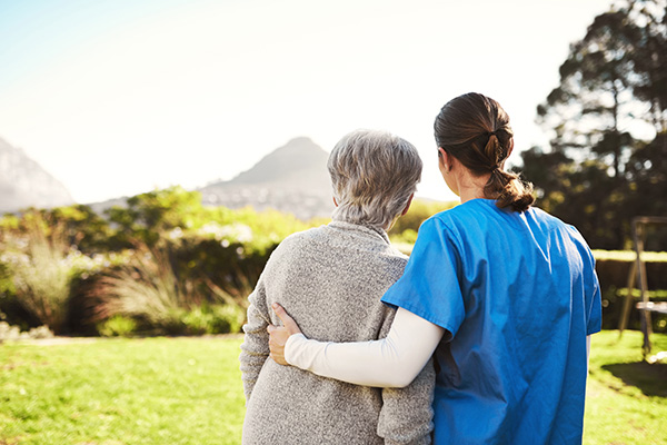 A healthcare worker and an elderly person stand outdoors, embracing and looking at the scenery.