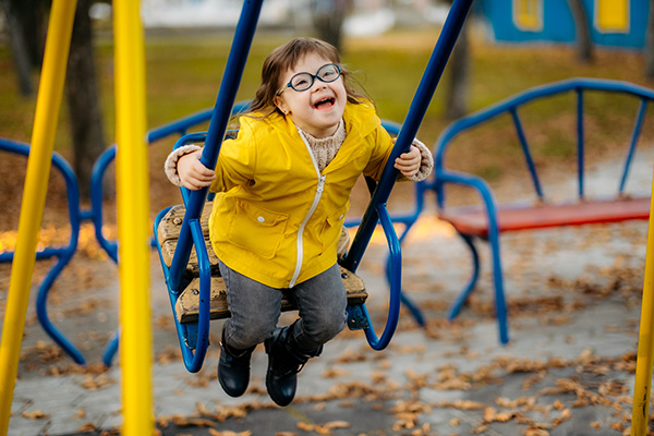 Child in a yellow jacket joyfully swings on a playground swing. Fallen leaves are scattered on the ground.