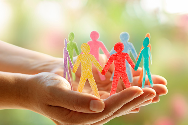 Hands holding cut-out paper figures of people in various colors, forming a circle against a blurred outdoor background.