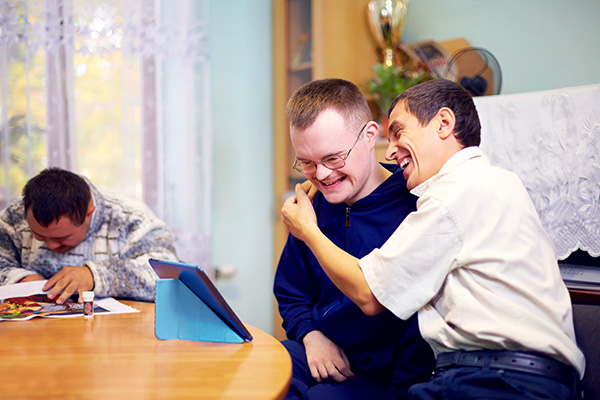 Three people are seated at a table; one is using a tablet, another is smiling, and the third is leaning over, playfully interacting with the one smiling.