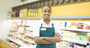 A store employee in a green apron stands smiling with arms crossed in front of refrigerated shelves stocked with various products.