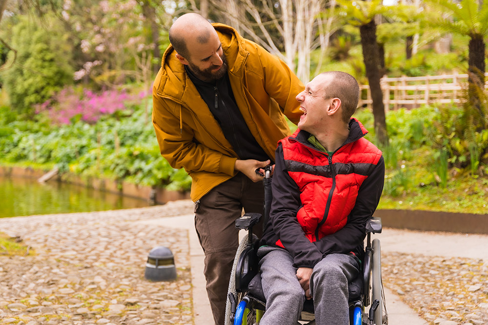 Two people are outdoors; one is pushing the other in a wheelchair. They are surrounded by greenery and cobblestones.