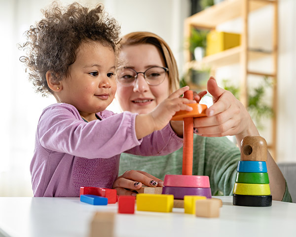 A child and adult play with colorful stacking toys on a table in a bright room.