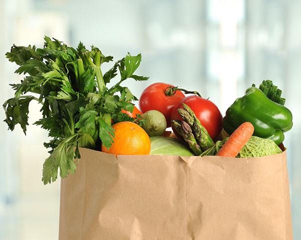 A paper bag filled with fresh vegetables and fruits, including celery, tomatoes, a green pepper, an orange, asparagus, a carrot, and a cabbage.