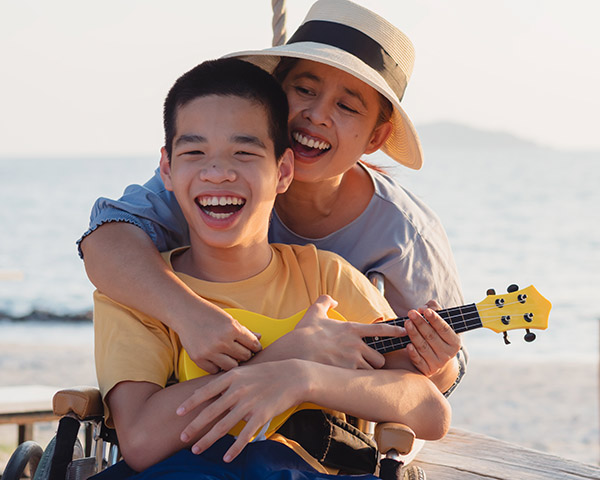 Child in a wheelchair holding a yellow ukulele, smiling, with a woman hugging him from behind, both enjoying a day at the beach.