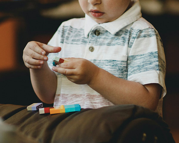 Child in a striped shirt is playing with building blocks, holding one block in each hand, with additional blocks on a surface in front.