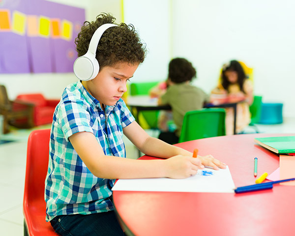 A boy wearing headphones is drawing with crayons at a red table in a classroom. Other children are sitting at tables in the background.