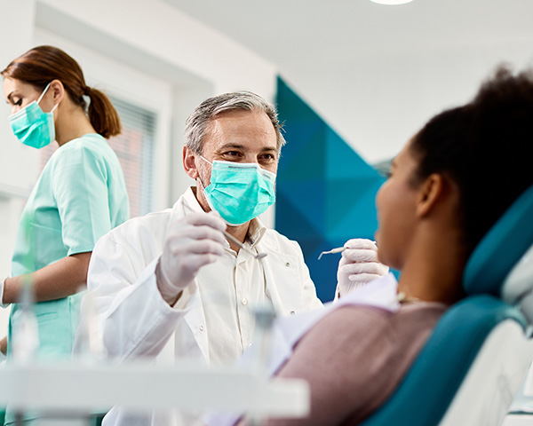 A dentist wearing a mask examines a patient in a dental chair, while an assistant prepares tools in the background.