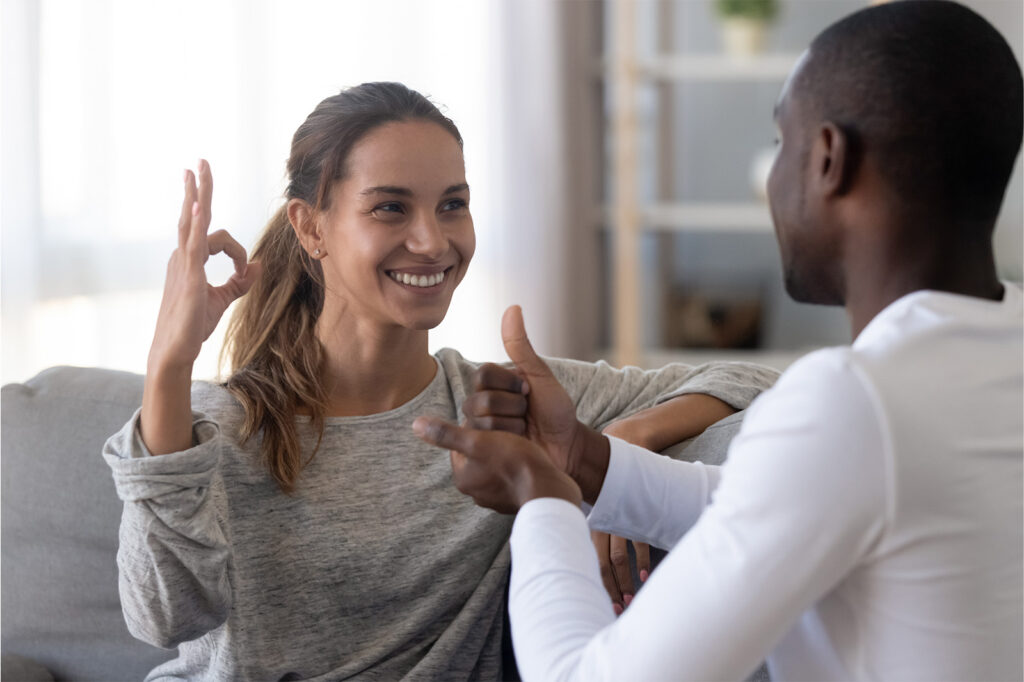 Two people sitting on a couch are engaging in a conversation using sign language. The woman is making an "OK" gesture, and the man is responding with a thumbs-up.