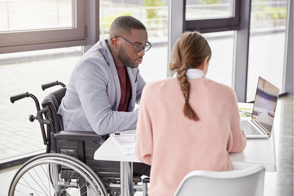 Man in a wheelchair and a woman sit at a table working on a laptop in a modern office setting.