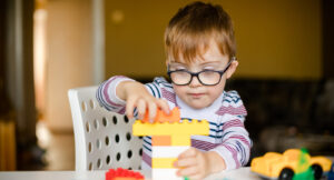 A young child with glasses builds a structure using colorful building blocks at a table.