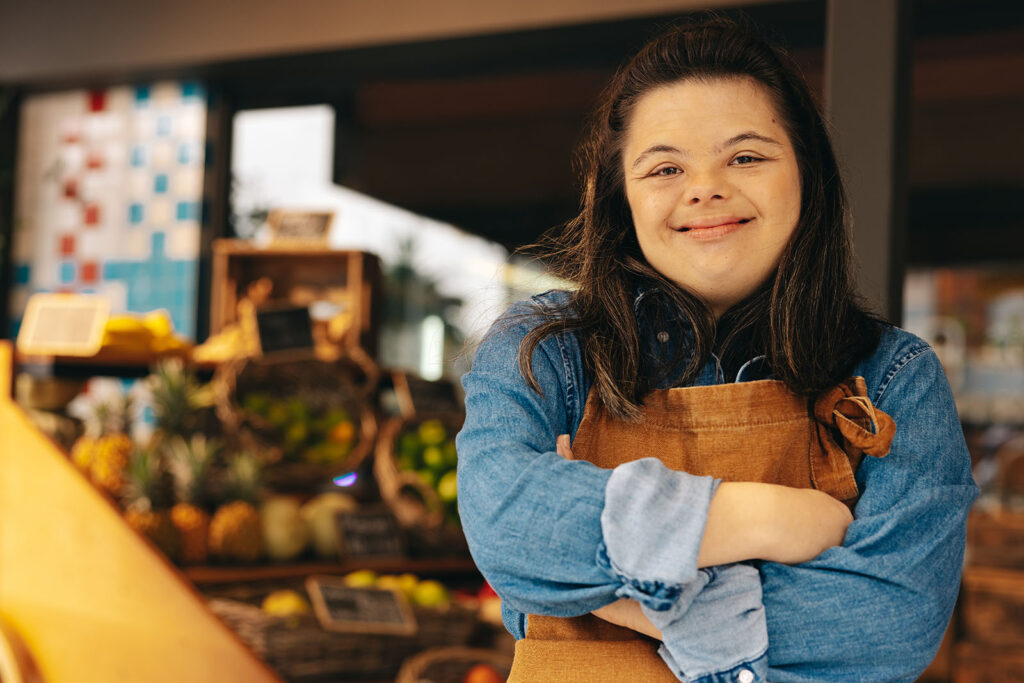 Person with long hair and an apron stands smiling with arms crossed in a market setting with fruit displayed in the background.