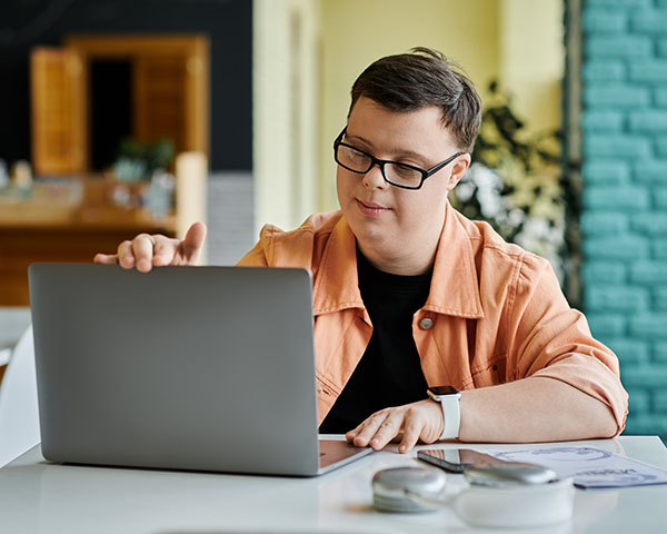 A person wearing glasses and an orange shirt types on a laptop at a desk with papers and a device nearby.