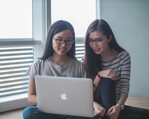 Two people sit on the floor by a window, looking at an open laptop together.