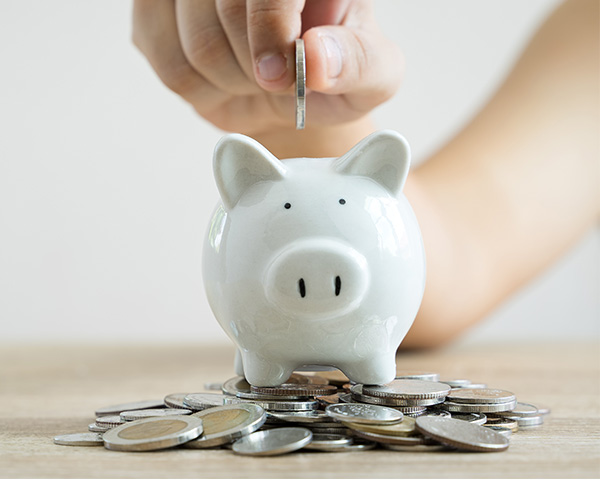 A hand inserts a coin into a white piggy bank on a wooden surface, surrounded by scattered coins.