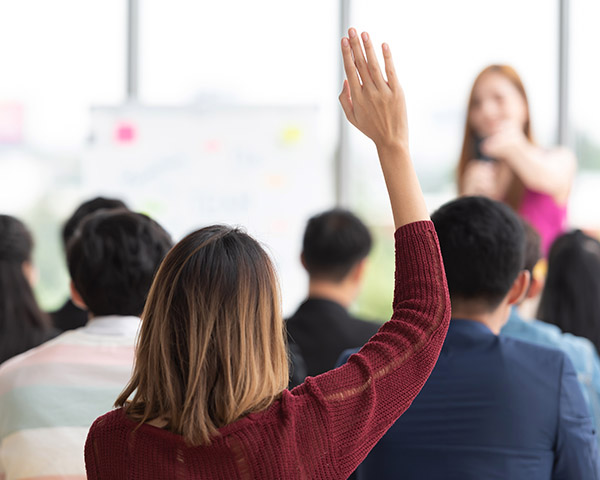 A woman raises her hand in a classroom setting, facing a speaker at the front, with a blurred whiteboard backdrop.