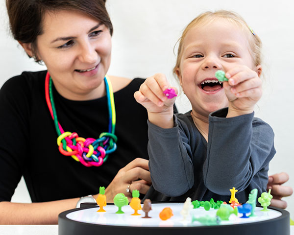 A woman and a child play with colorful toy figures on a light table. The child is smiling and holding two toys.