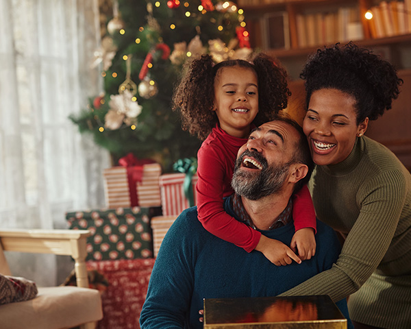 A family of three smiles and embraces by a Christmas tree, with wrapped gifts in the background.