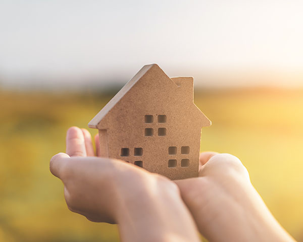 Hands holding a small wooden house model against a blurred outdoor background.