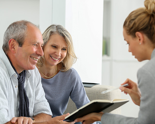 A couple sitting on a couch listens to a businesswoman holding a notebook and a pen.