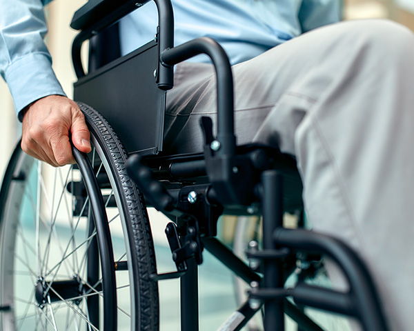 Person in a wheelchair, hand on the wheel, wearing a light blue shirt and beige pants.