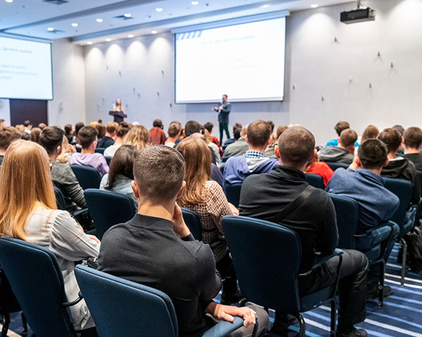 Audience attending a presentation in a conference room, with one speaker at the podium and another standing on stage, in front of two projection screens.