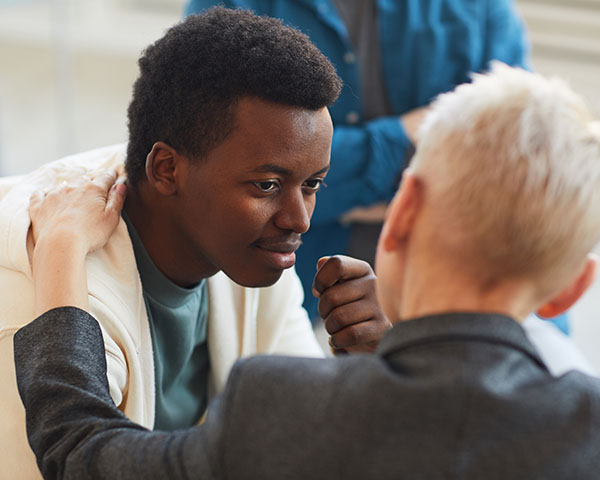 A person with short dark hair is seated and listening intently to another person with short blond hair who is speaking to them, placing a hand on their shoulder in a supportive gesture.