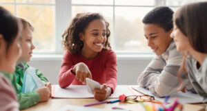 A group of children sitting at a table, talking and smiling, with notebooks and school supplies in front of them.