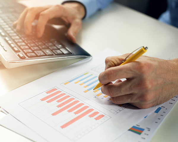 Person analyzing printed graphs with a yellow pencil while typing on a keyboard.