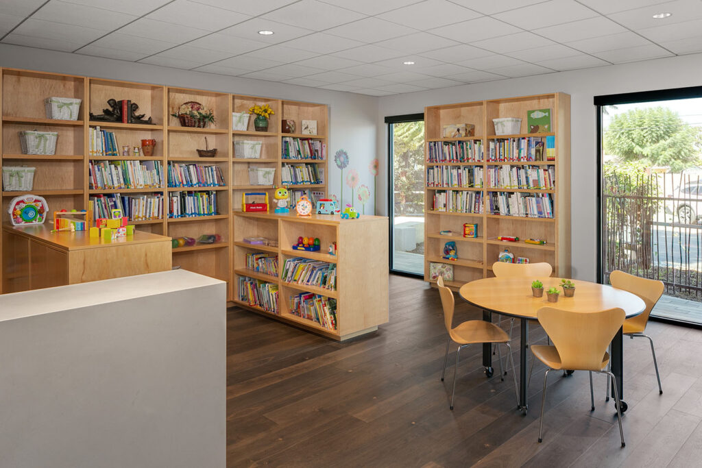 A cozy library corner with wooden shelves filled with books and toys, a round table with chairs, and windows letting in natural light.