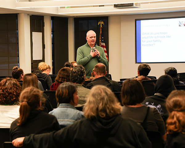 A man stands speaking into a microphone in a room full of seated people facing a presentation screen.