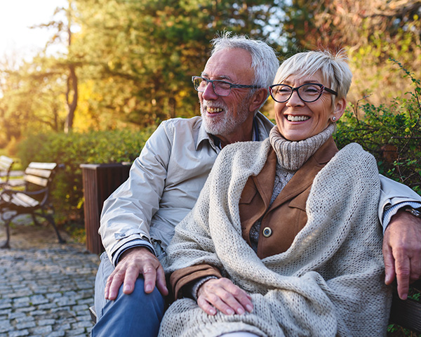 Elderly couple sitting on a park bench, smiling and wrapped in a blanket, surrounded by trees and autumn foliage.