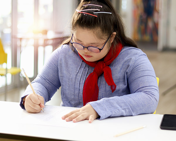 A girl with glasses and a red scarf writes in a notebook at a table in a bright room.