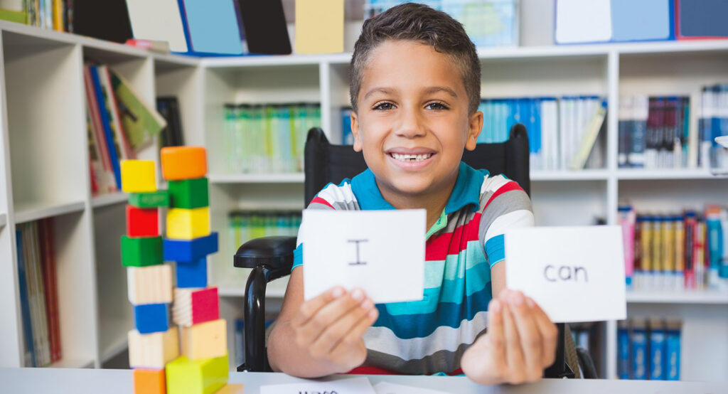 A smiling boy holds up two cards reading "I" and "can" in a library. Colorful wooden blocks are stacked on a table beside him.