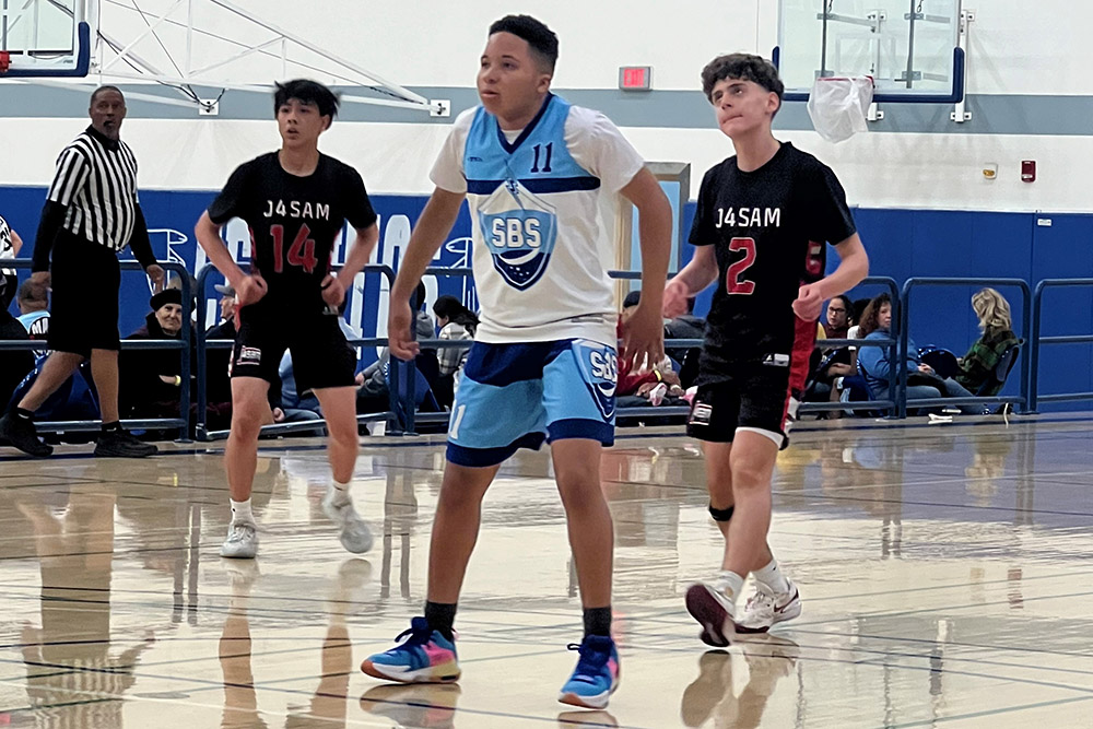 Three basketball players stand on an indoor court ready for action. One wears a blue and white jersey, and the other two wear black jerseys. Spectators are seated in the background.