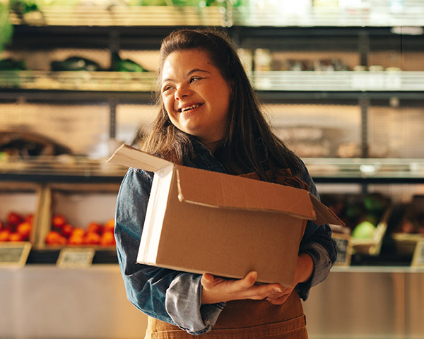 A person smiling while holding a cardboard box in a grocery store, with shelves of produce in the background.