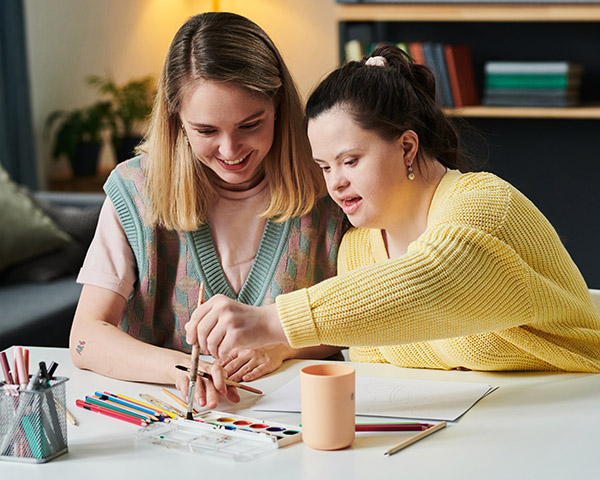 Two people are sitting at a table, engaging in a joint art activity with colored pencils and paint. They appear focused and collaborative.