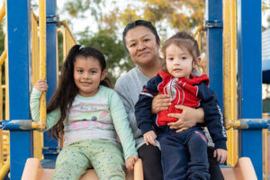 A woman sits on a playground slide holding a toddler, with a young girl sitting next to them. There are trees in the background.