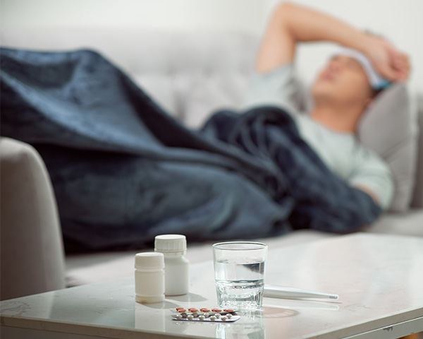 Person lying on a couch under a blanket with a thermometer, medicine bottles, and a glass of water on the table.