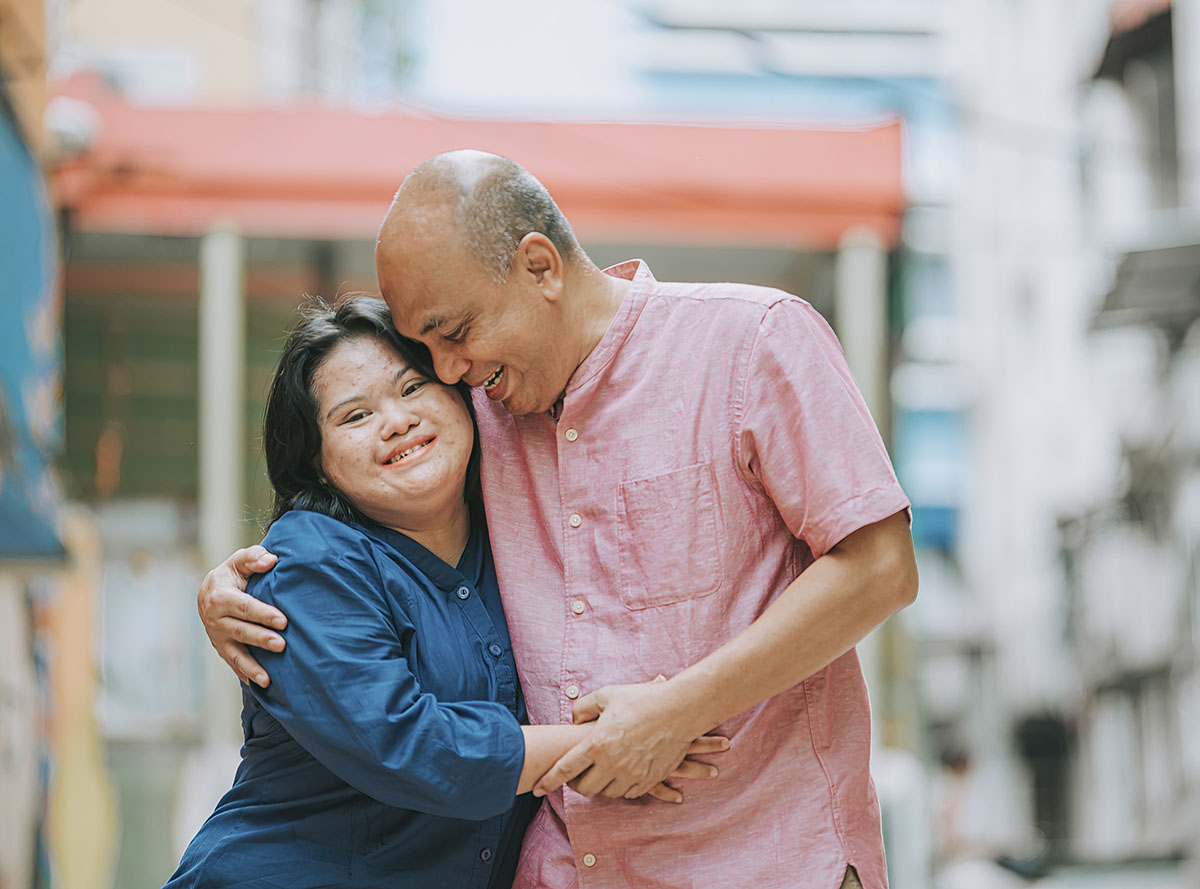 Một người đàn ông và một người phụ nữ đang ôm nhau ấm áp trên phố. Người đàn ông mặc áo sơ mi màu hồng, và người phụ nữ mặc áo sơ mi màu xanh. Họ đang mỉm cười và có vẻ hạnh phúc.