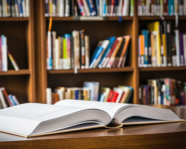 Open book on a wooden table in a library, with shelves of blurred books in the background.