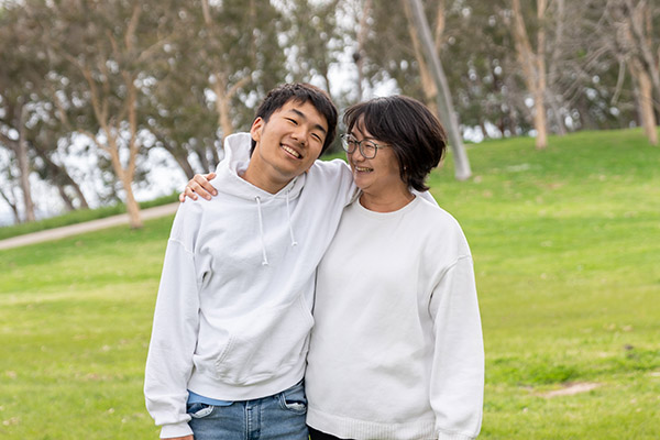 A person with short hair and another with glasses and shoulder-length hair stand together smiling in a green park. They wear white tops.