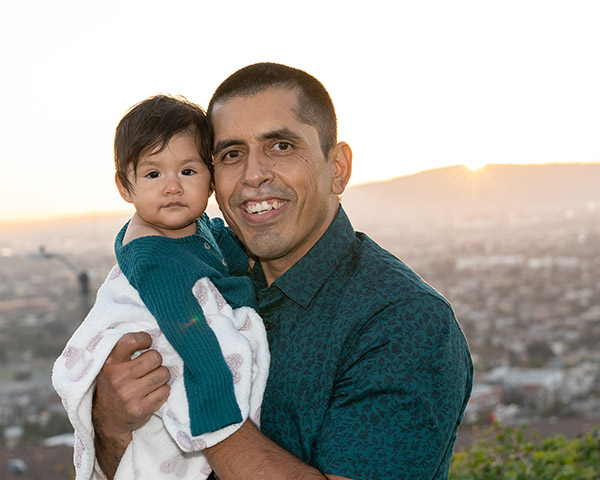 A man holds a baby wrapped in a blanket, with a cityscape and sunset in the background.